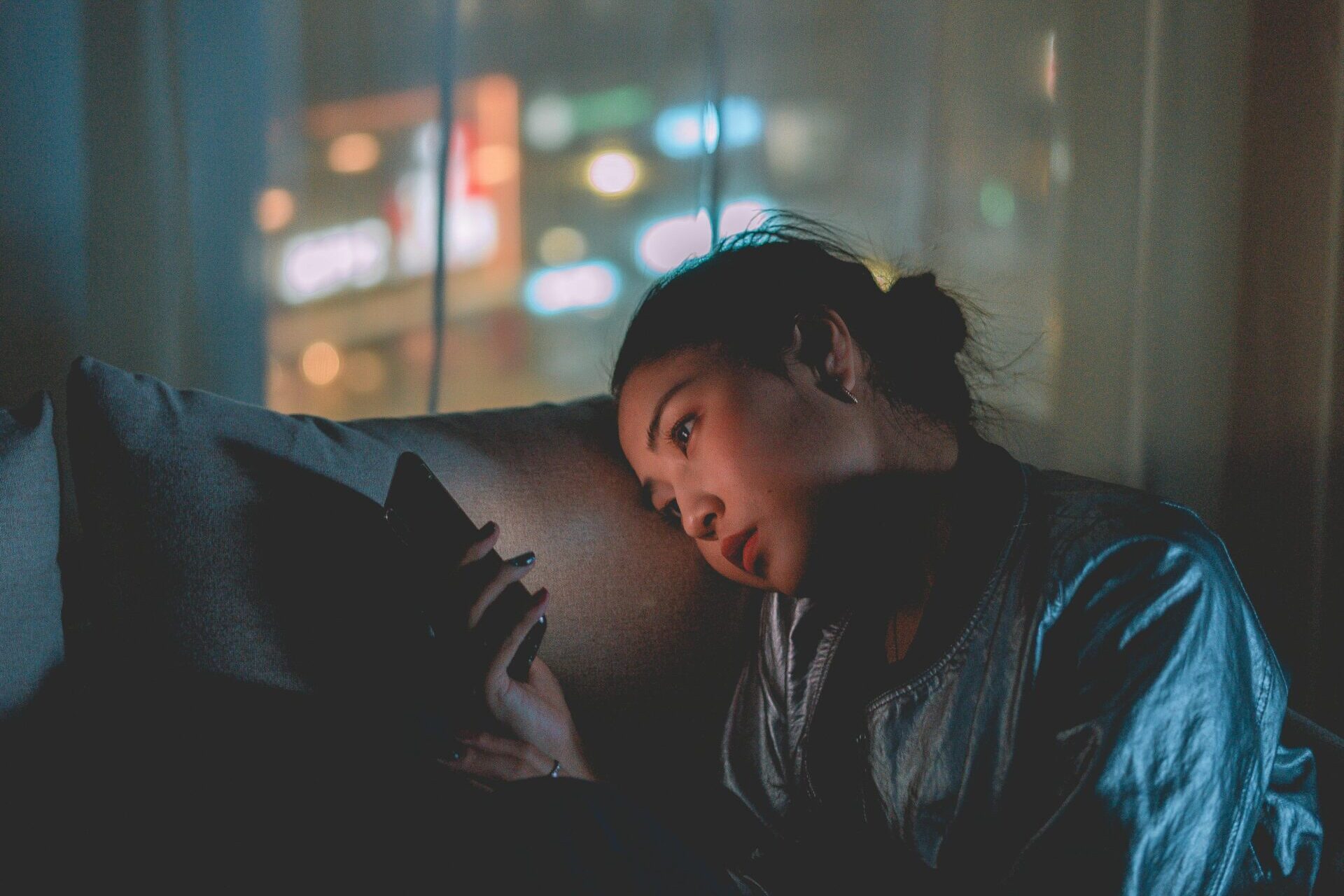 A woman looking at her mobile phone at night with city lights blurred in the background.